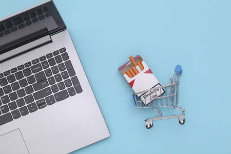 Laptop and shopping trolley with pack of cigarettes on blue background. top viewの写真素材