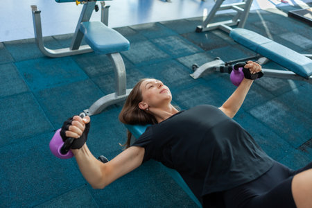 Young fitness woman trains pectoral muscles with a kettlebells in her hands while lying on a bench...
