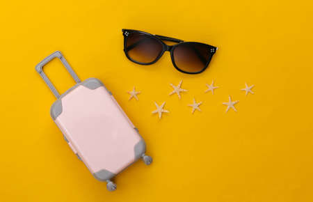 Travel or beach resort flat lay. Mini plastic travel suitcase, sunglasses and starfish on yellow background. minimal style. top viewの写真素材