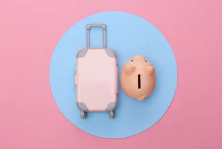 Travel minimalism. Mini plastic travel suitcase and piggy bank on pink background with blue circle. minimal style. Top view, flat layの写真素材