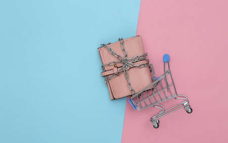 Mini shopping trolley with leather wallet wrapped in steel chain on pink blue background. top view