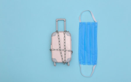 Mini travel baggage wrapped in steel chain and medical mask on blue background.