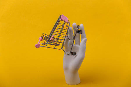 White mannequin hand holding mini shopping trolley on yellow background.の写真素材