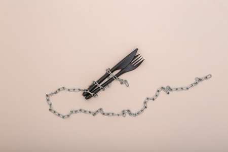 Plastic fork and knife wrapped in a steel chain on beige background. Diet conceptの写真素材