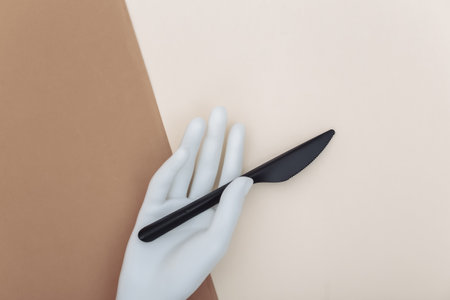 White mannequin hand holds a plastic knife on beige brown background. minimalism. top viewの写真素材