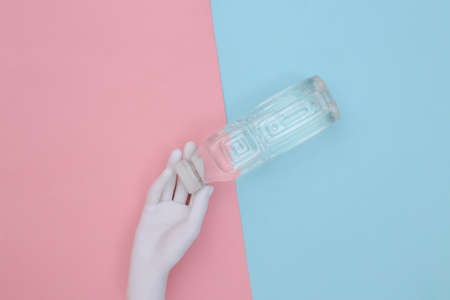 White mannequin hand holding a bottle of water on a blue-pink pastel background. top view....