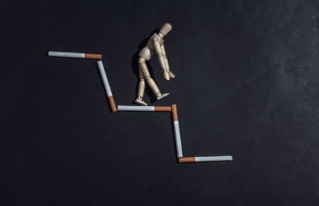 Social degradation due to bad habits. Oppressed wooden puppet steps down stairs from cigarettes on black backgroundの写真素材