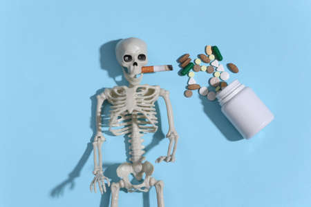 smoking cure. Smoking skeleton with a cigarette and a bottle of pills on blue background. Quitting smokingの写真素材