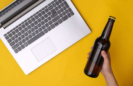 Hand hold a bottle of beer on a yellow background with a laptop. The concept of rest, relaxation after a hard day's work. top viewの写真素材