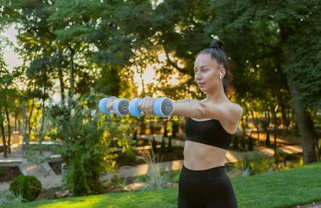 Attractive caucasian fit brunette woman training muscles with dumbbells in her hands in the park at...