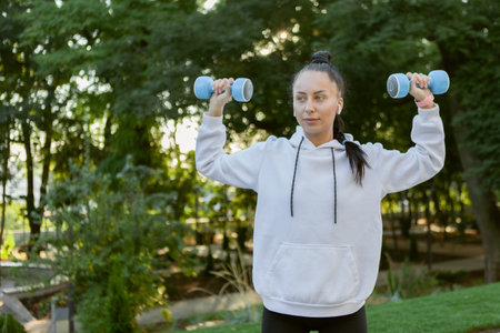 Attractive caucasian fit brunette woman training muscles with dumbbells in her hands in the park at sunriseの写真素材