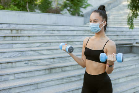 Young slim woman in a medical protective mask trains with dumbbells in her hands outdoors. Sports during covid-19 periodの写真素材