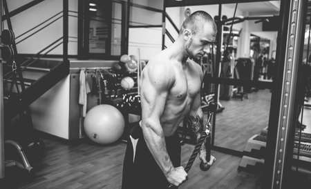 Muscular bodybuilder handsome man with naked torso trains triceps in a crossover cable exercise...