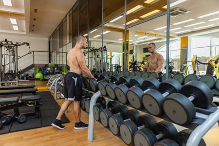 Muscular man with a naked torso removes dumbbells from a rack while looking in a mirror in a modern...