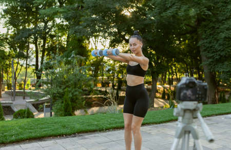 Online fitness trainer. Young attractive fit woman shows how to do exercise with dumbbells, filming blog on camera outdoors in the morning. Sports blog, fitness vlogの写真素材