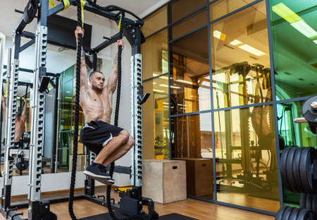 Young muscular fitness man training abdominal muscles while hanging on the ropes in gym