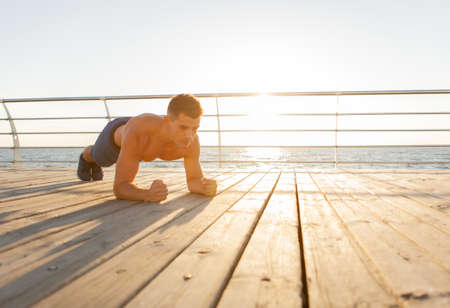 Young muscular man trains endurance and practicing plank exercise at sunrise on the beachの写真素材
