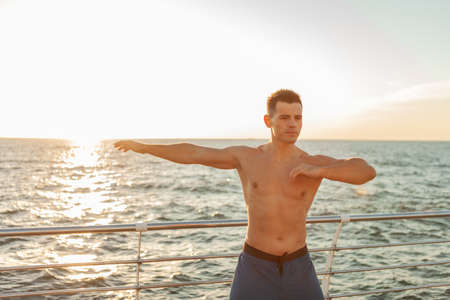 Muscular handsome man with naked torso doing warm up exercise before workout on the beach at sunrise