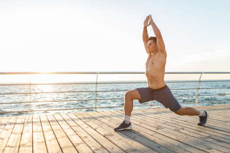 Muscular handsome man with a naked torso practices the warrior 1 pose asana on the beach at sunrise....