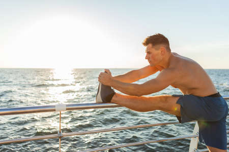 Young muscular man with naked torso doing leg stretching on the beach at sunrise