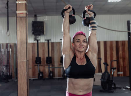 Muscular extraordinary female athlete with short pink hair trains overhead with heavy kettlebells. Functional, cross training in modern gym.の写真素材