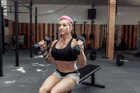 Muscular athletic woman trains biceps with dumbbells in her hands while sitting on a bench in the gym.の写真素材