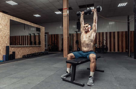 Powerful athletic man training shoulder muscles with heavy dumbbells doing overhead while sitting on...