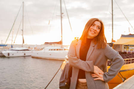 Portrait of a young cheerful woman in an autumn coat posing in a yacht club at sunriseの写真素材