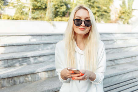 Young attractive blonde woman in sunglasses using smartphone while sitting on a bench in city parkの写真素材