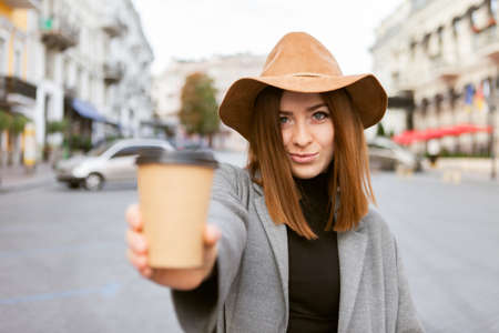 Stylish millennial woman in a slicker coat and felt hat jerks a coffee cup on the go while walking in a european cityの写真素材