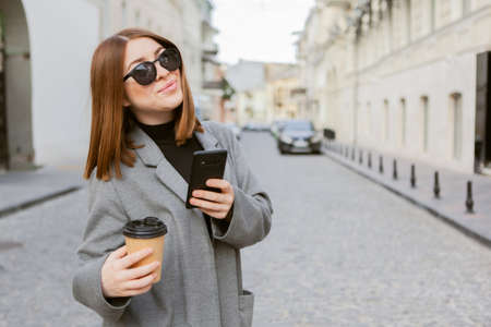 Young millennial smiling woman with smooth hair dressed in an autumn coat and sunglasses uses a smartphone and holds a cup of coffee on the go in a European city.の写真素材