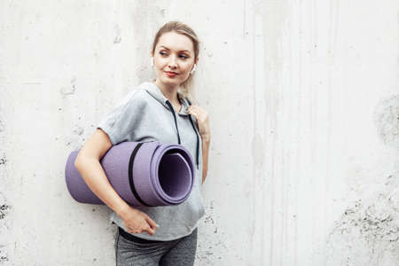 Fitness sport girl in fashion sportswear holding rolled mat against concrete wall, outdoor sports, urban styleの写真素材