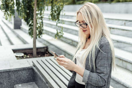 Attractive business woman holding smartphone in her hands while sitting on bench outdoorsの写真素材
