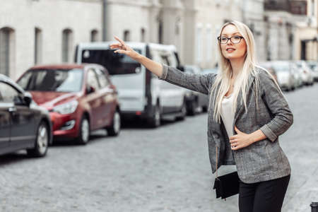 Beautiful Woman Catching Taxi Car On Street. Portrait Blonde Business Woman On Way To Work Stopping Taxi Car Outdoors.の写真素材