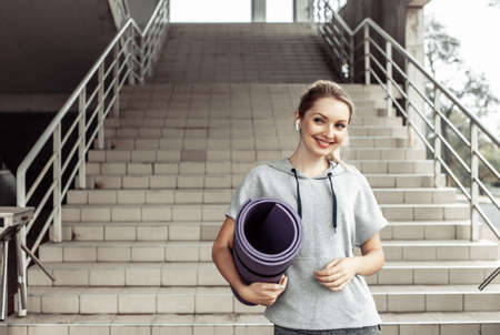 Fitness sport woman in fashion sportswear and wireless earphones holding rolled mat against stairs,...