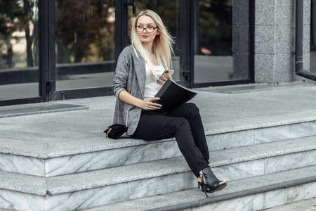 Busy business woman sitting on the steps during the break and holding folder and smartphone on the background of a business centerの写真素材