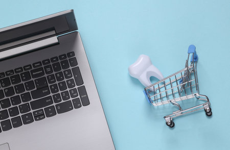 Fake tooth in shopping trolley and laptop on blue background. Teeth care, medicine concept. top view