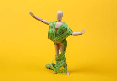 Diet concept, weight loss. Wooden puppet wrapped with measuring tape isolated on yellow background
