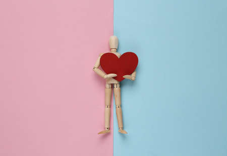 Wooden puppet mannequin holding red heart on pink blue pastel background.