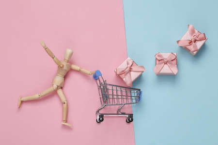 Wooden puppet rolls supermarket trolley with gift boxes on pink blue pastel background. Holiday shopping concept.の写真素材