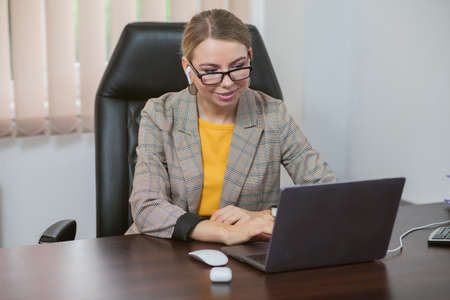 Young cheerful business woman with wireless headphones sits at a table with a laptop in her officeの写真素材