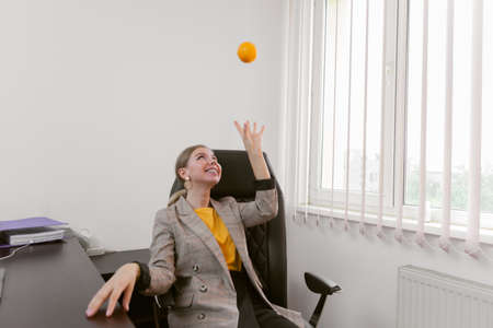 Cheerful business lady tossing an orange while sitting at the table in her officeの写真素材
