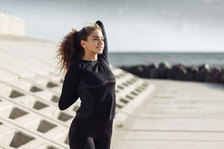 Outdoor workout concept. Fit slim woman with curly hair stretches or warms up on an urban embankment...