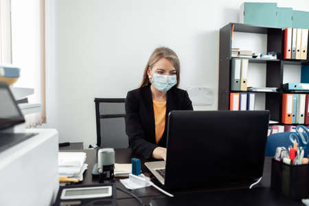 Business woman in medical mask working on a laptop in her officeの写真素材