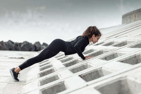 Outdoor workout concept. Fit slim woman with curly hair does push-ups on an urban embankment with concrete slabs. healthy lifestyleの写真素材