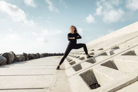 Outdoor workout concept. Fit slim woman with curly hair stretches or warms up on an urban embankment with concrete slabsの写真素材