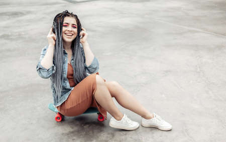Extraordinary female millennial skater with colorful makeup, African braids sits on penny board and...