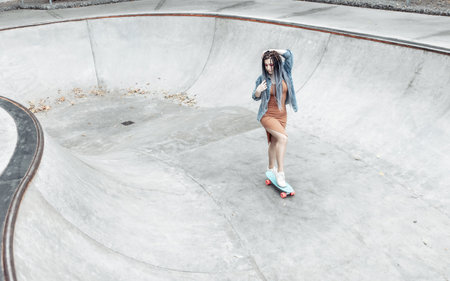Cheerful extraordinary millennial woman with bright makeup and African braids is riding a penny board in a skateparkの写真素材