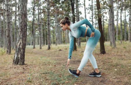 Attractive fitness woman with perfect body doing warm-up or stretching exercise in the forest....