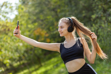 Smiling slim fit blonde woman in headphones makes selfie on smartphone in park